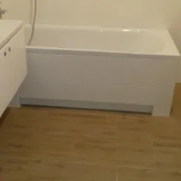 Nowoczesna łazienka z białą wanną obudowaną płytkami, umywalką z szafką i prysznicem, wykończona płytkami imitującymi drewno na podłodze.