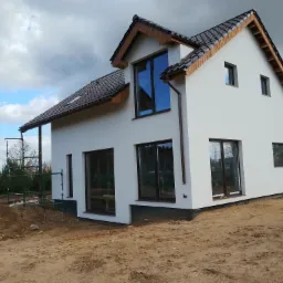Nowoczesny, biały dom z brązowym dachem i ramami okiennymi, widoczny z zewnątrz na etapie wykończeniowym, z widocznymi rusztowaniami i przygotowanym terenem.