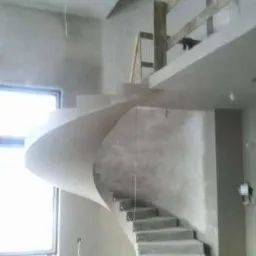 Surowe, spiralne schody z tynkowanymi ścianami w trakcie wykańczania wnętrza, widoczne przewody elektryczne i okno.