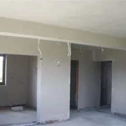 Wnętrze domu po nałożeniu świeżych, jasnoszarych tynków maszynowych; widoczne kable elektryczne wystające ze ścian i sufitu, przygotowane do dalszych prac instalacyjnych; w tle okno i worki...