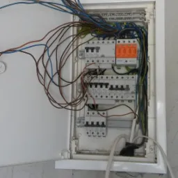 Otwarta skrzynka bezpiecznikowa z widocznymi przewodami i elementami zabezpieczeń na szarej, surowej ścianie.