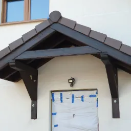 Zadaszenie nad wejściem do budynku, wykonane z ciemnego drewna i brązowej dachówki, z zabezpieczonym folią i taśmą wejściem, oświetlone lampą z czujnikiem ruchu.