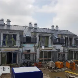 Wielorodzinny budynek w trakcie budowy, z szarą blachą na dachu, widoczne kominy, ocieplenie ścian styropianem i materiały budowlane na błotnistym placu budowy.