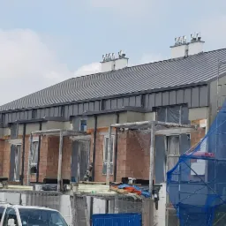 Szeregówka w trakcie budowy z szarym, metalowym pokryciem dachowym i kominami, widoczne rusztowania i materiały budowlane.