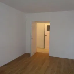 Pomalowane na biało wnętrze pokoju z drewnianą podłogą i otwartymi białymi drzwiami do następnego pomieszczenia, gdzie widać kolejne białe drzwi.