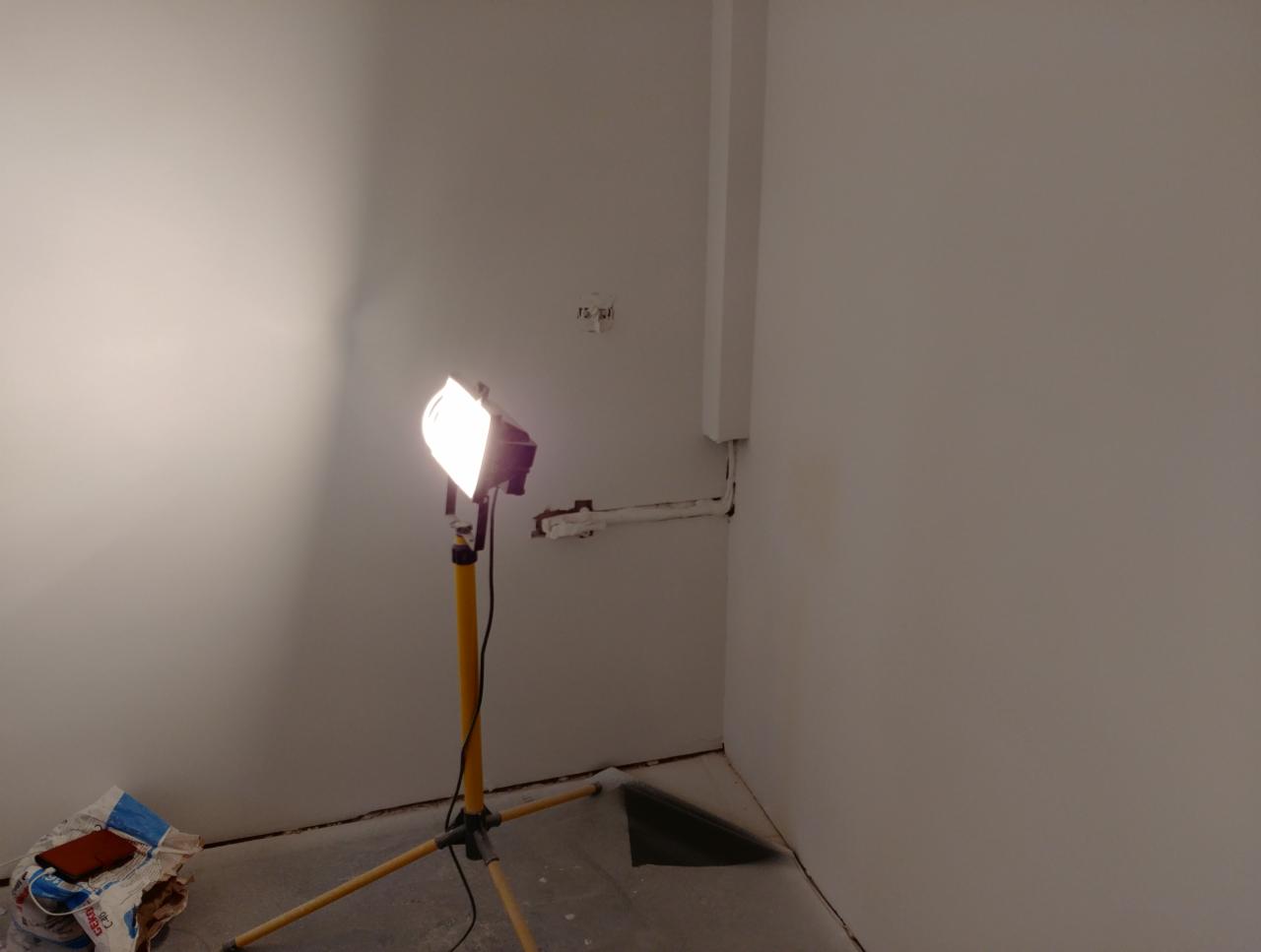 Świeżo pomalowane, białe ściany w rogu pokoju oświetlone intensywnym światłem halogenowym na żółtym statywie. Widoczne ślady po instalacji elektrycznej i resztki materiałów budowlanych w worku.