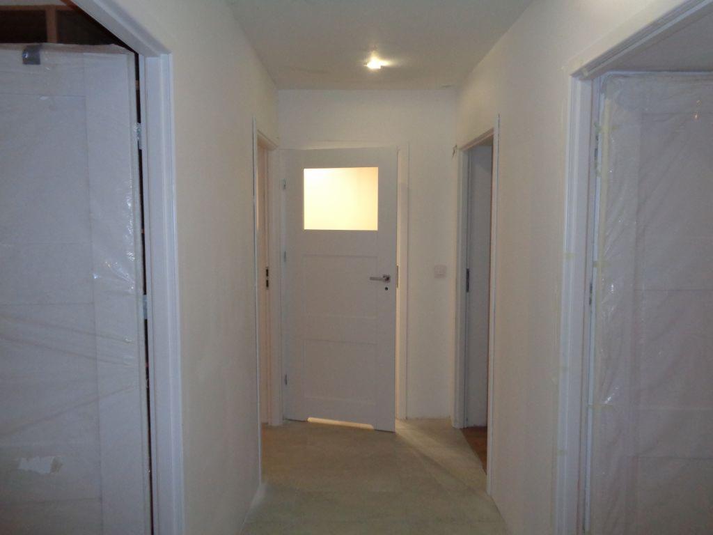 Korytarz w trakcie odświeżania: ściany pomalowane na biało, nowe białe drzwi częściowo zabezpieczone folią ochronną, widoczne przygotowania do dalszych prac wykończeniowych.