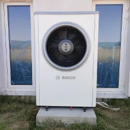 Biała jednostka zewnętrzna pompy ciepła marki Bosch, umieszczona na betonowym fundamencie przed ścianą budynku z dwoma oknami zabezpieczonymi folią.