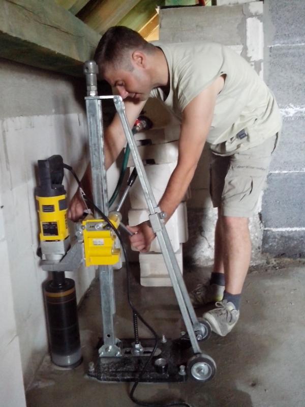 Hydraulik wierci otwór w ścianie za pomocą wiertnicy diamentowej z odsysaniem wody, wewnątrz surowego budynku z betonowymi ścianami.
