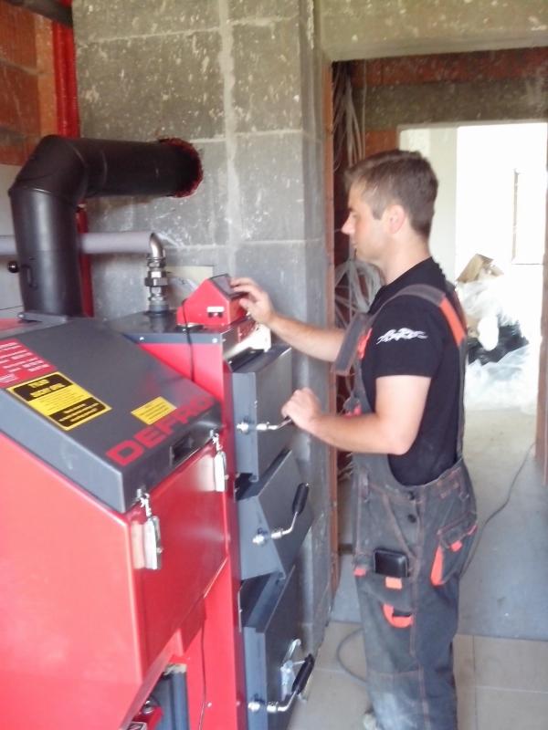 Instalator w kombinezonie obsługuje czerwono-szary piec Defro w surowym wnętrzu budynku w trakcie budowy.