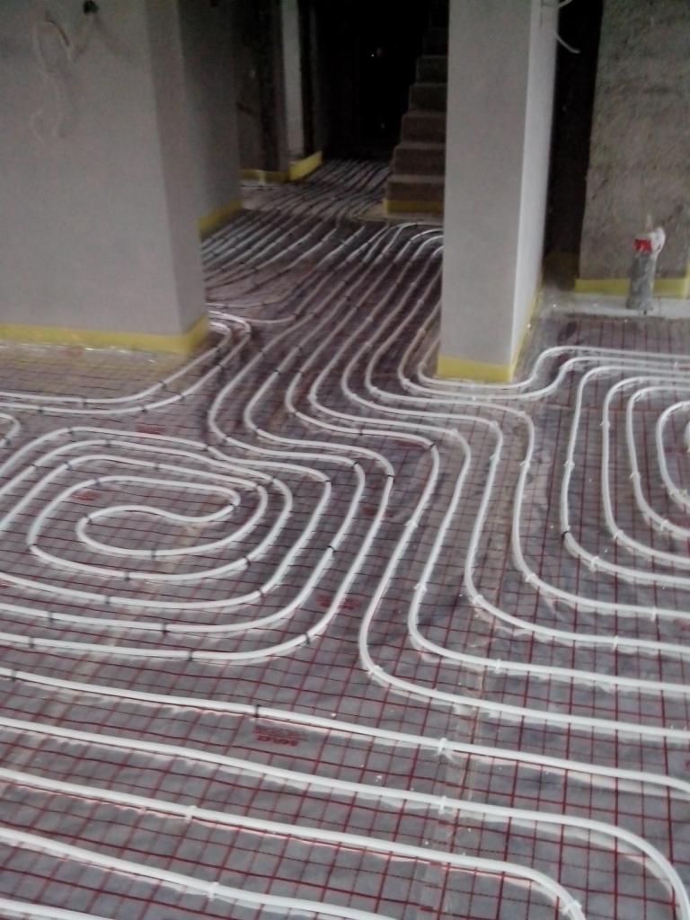 Białe rurki ogrzewania podłogowego ułożone na siatce, widok z góry, surowy stan wykończenia wnętrza, żółta taśma na ścianach.