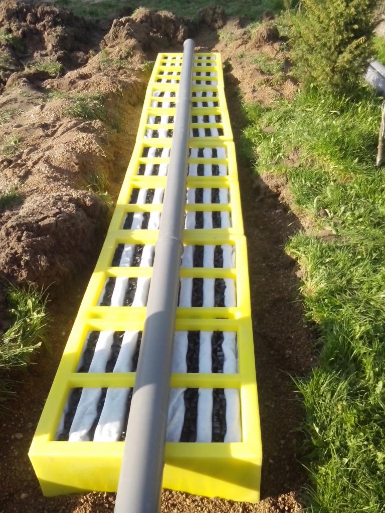 Żółta skrzynka rozsączająca z rurą odprowadzającą, ułożona w wykopie ziemnym na trawniku.