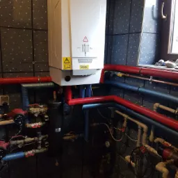 Biały piec gazowy De Dietrich z czerwonymi i niebieskimi rurami na tle ciemnych płytek w Werbkowicach, widoczny regulator ciśnienia i okno w tle.