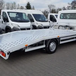 Biała autolaweta z aluminiową platformą, widoczna z boku, na tle białych busów zaparkowanych na szarym brukowanym placu.