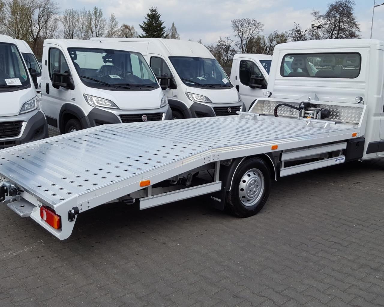 Biała autolaweta z aluminiową platformą, widoczna z boku, na tle białych busów zaparkowanych na szarym brukowanym placu.