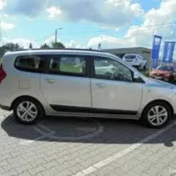 Srebrny, rodzinny van na kostce brukowej, zaparkowany na tle błękitnego nieba z chmurami i flagami reklamowymi w tle. Widoczny z boku, lekko od tyłu.