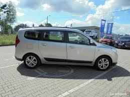 Srebrny, rodzinny van na kostce brukowej, zaparkowany na tle błękitnego nieba z chmurami i flagami reklamowymi w tle. Widoczny z boku, lekko od tyłu.