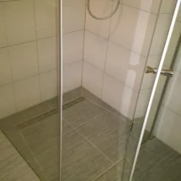 Nowoczesna kabina prysznicowa z odpływem liniowym, wykończona płytkami w odcieniach szarości, z chromowaną armaturą i szklanymi drzwiami.