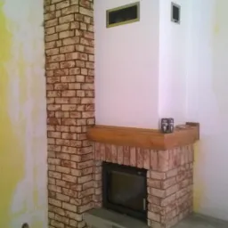 Wykończony kominek z cegły z drewnianą półką w rogu pokoju, widoczne niedomalowane ściany i surowa podłoga.