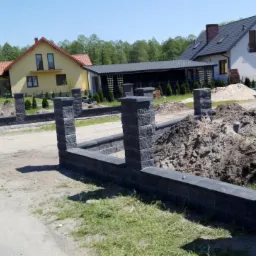 Wznoszenie ogrodzenia z bloczków betonowych w kolorze grafitowym, widoczne słupki i podmurówka, w tle dwa domy jednorodzinne i drzewa.
