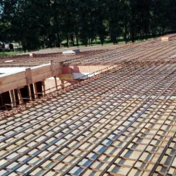 Siatka zbrojeniowa ułożona na stropie budynku w trakcie budowy, widoczne szalunki i pustaki ceramiczne, tło stanowią drzewa.