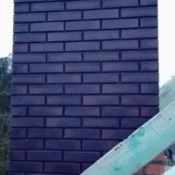 Murowany komin z ciemnej cegły klinkierowej, z widocznymi otworami wentylacyjnymi, na tle konstrukcji dachu z surowego drewna.