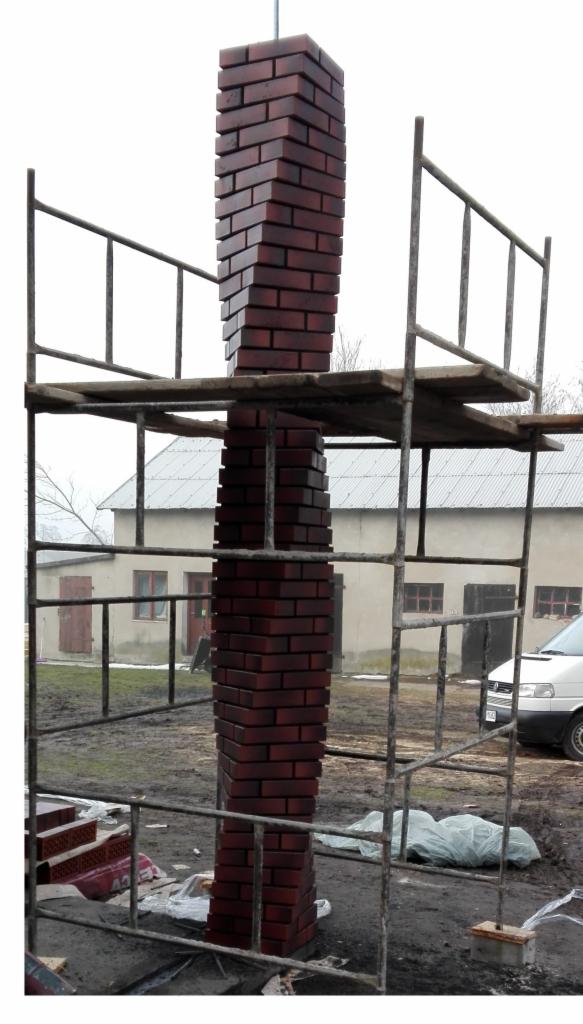 Wznoszona z czerwonej cegły kolumna, otoczona rusztowaniem, na tle budynku gospodarczego i pochmurnego nieba.