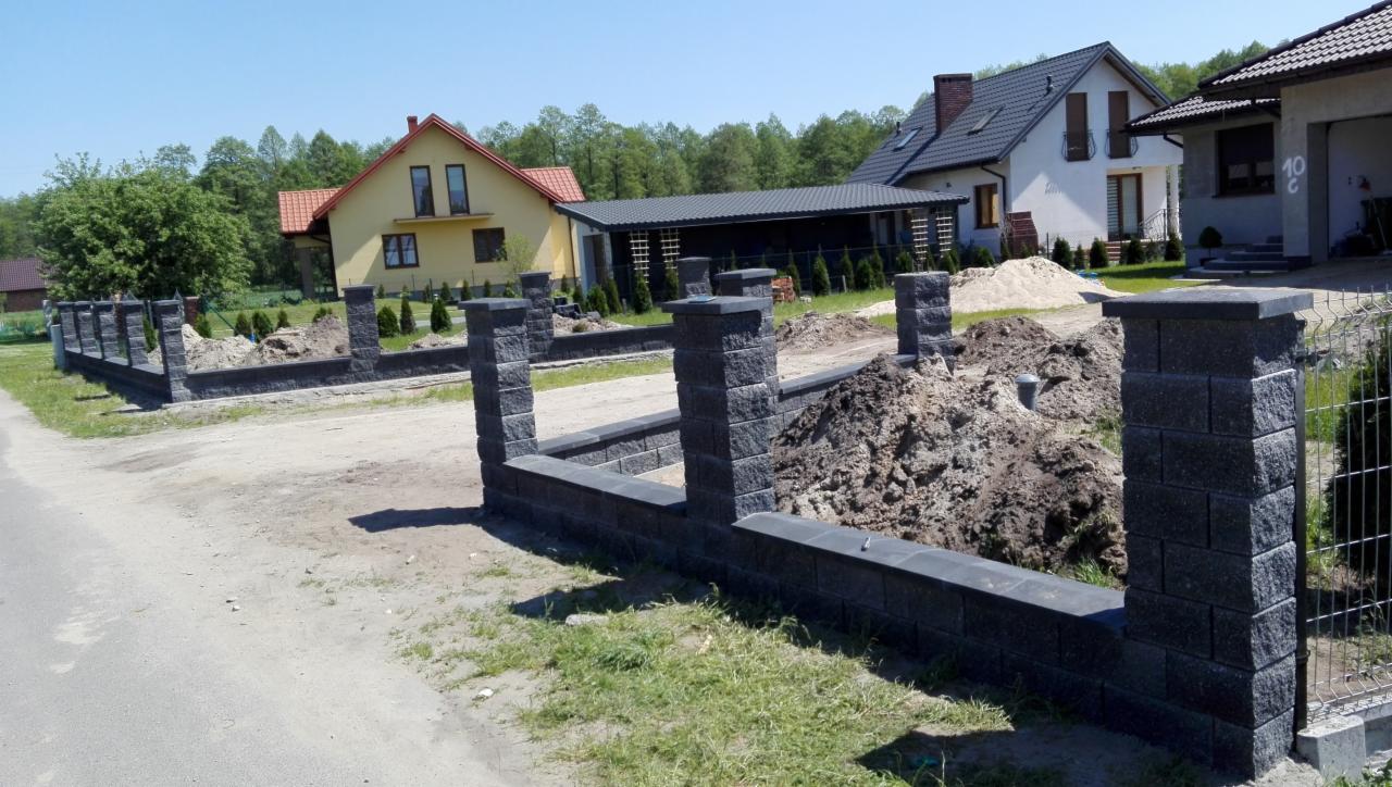 Wznoszenie ogrodzenia z bloczków betonowych w kolorze grafitowym, widoczne słupki i podmurówka, w tle dwa domy jednorodzinne i drzewa.