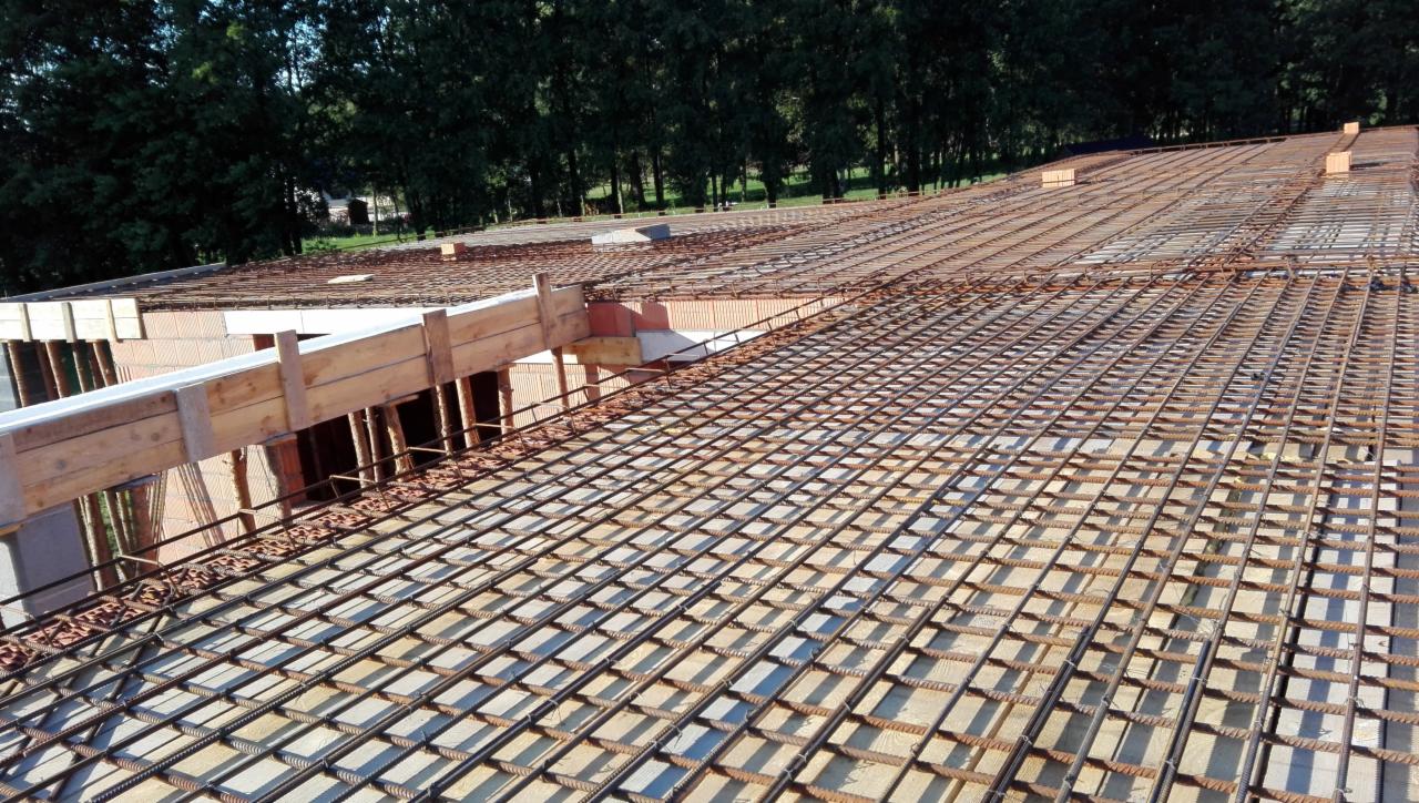 Siatka zbrojeniowa ułożona na stropie budynku w trakcie budowy, widoczne szalunki i pustaki ceramiczne, tło stanowią drzewa.