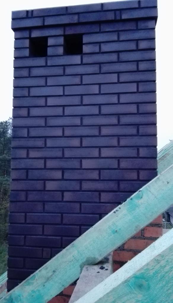 Murowany komin z ciemnej cegły klinkierowej, z widocznymi otworami wentylacyjnymi, na tle konstrukcji dachu z surowego drewna.
