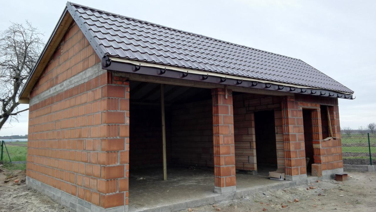 Surowy budynek z czerwonej cegły z brązowym dachem z blachodachówki i zamontowanymi rynnami, widok z zewnątrz na otwartą przestrzeń wewnątrz budynku, częściowo wykończony fundament.