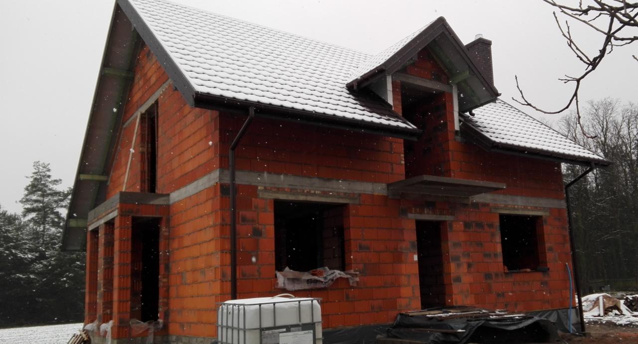 Niedokończony budynek z czerwonej cegły ceramicznej w zimowy dzień, z widoczną konstrukcją dachu pokrytą śniegiem i pustymi otworami okiennymi, na pierwszym planie biały zbiornik na wodę.