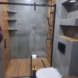 Nowoczesna łazienka z kabiną prysznicową typu walk-in, szare płytki ścienne z wnękami, drewnopodobne płytki podłogowe w strefie prysznica, wisząca toaleta i czarne akcenty.