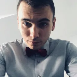 Portret mężczyzny z bordową muchą, w szarej koszuli, patrzącego z góry w dół, z widocznym oświetleniem padającym z góry. Ujęcie bliskie, skupione na twarzy i górnej części ciała.