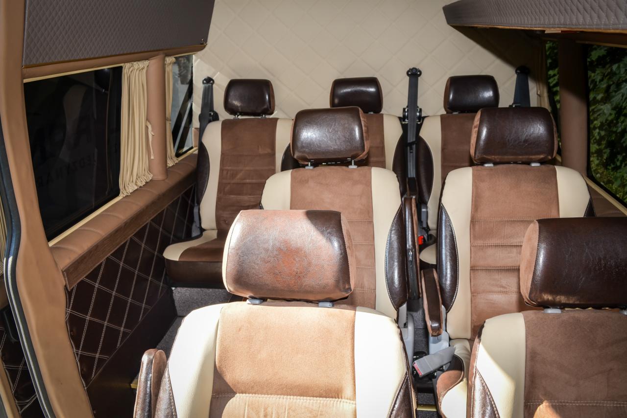 Wnętrze luksusowo wykończonego busa z beżowo-brązowymi skórzanymi fotelami, pikowaną tapicerką ścian i zasłonkami w oknach.