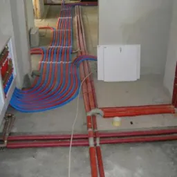 Rozbudowana instalacja grzewcza i wodna w surowym stanie, widoczne rury w kolorach czerwonym i niebieskim, rozdzielacz w białej obudowie oraz fragment ściany z białym włazem rewizyjnym.