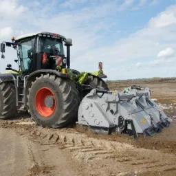 Ciągnik rolniczy z frezem glebowym podczas rekultywacji terenu, widoczne ślady pracy i przygotowywana gleba pod uprawy, w tle turbiny wiatrowe.