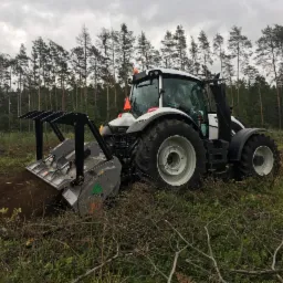 Biało-szary ciągnik z zamontowanym mulczerem leśnym, pracujący na zalesionym terenie, widoczne świeżo zmielone gałęzie i ścięte krzewy, pochmurne niebo w tle.