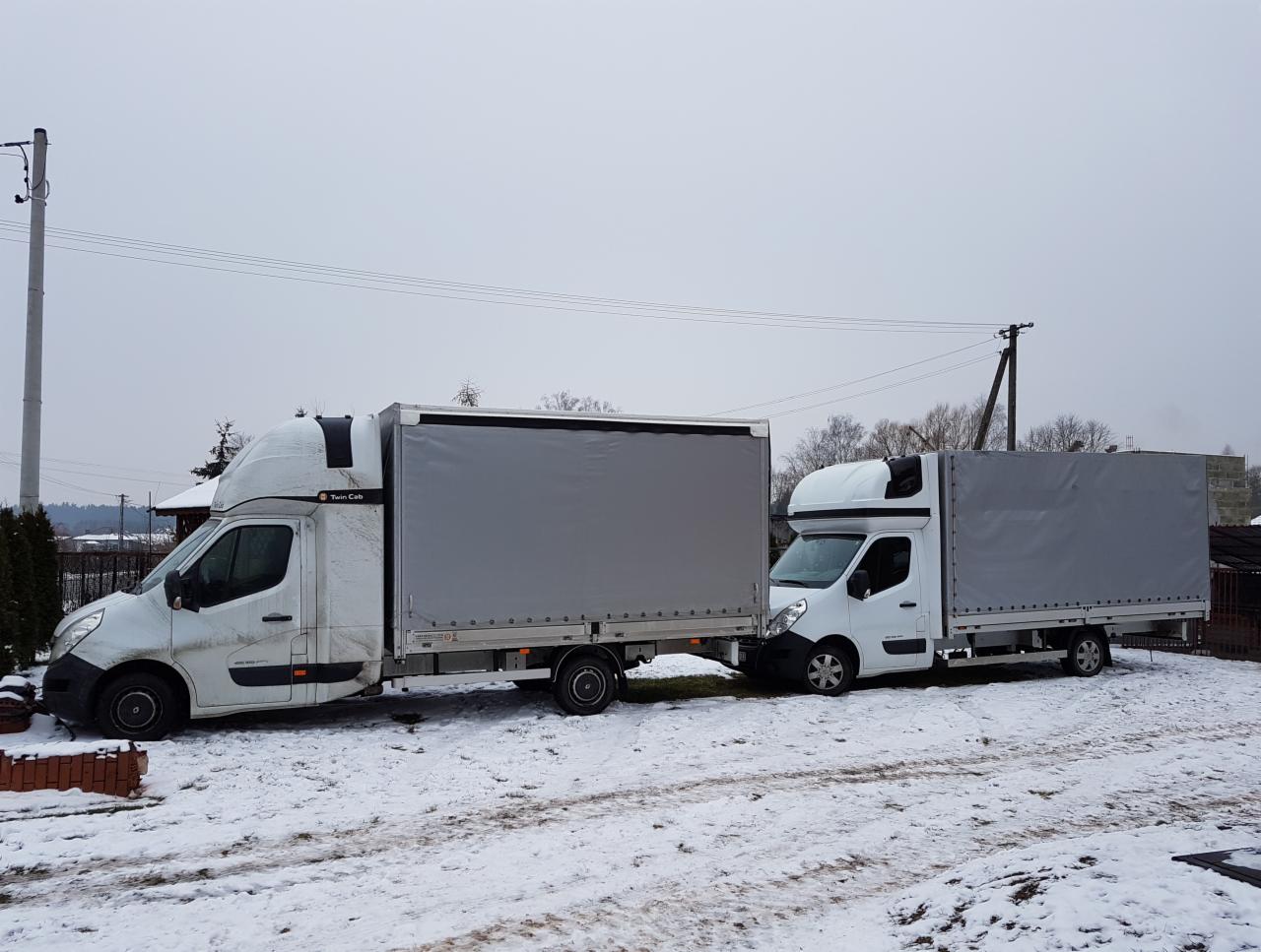 Dwa białe busy dostawcze z szarymi plandekami, zaparkowane obok siebie na zaśnieżonym terenie w Białobrzegach, widoczne przewody elektryczne i słupy w tle.