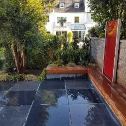 Nowoczesny taras z ciemnych płyt, drewnianą ławką z elementami z betonu architektonicznego, dekoracyjnym panelem w kolorze czerwonym i zielenią w tle.