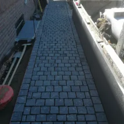 Ułożona ścieżka z ciemnej kostki granitowej pomiędzy budynkiem z cegły a betonowym korytem, widoczne narzędzia i materiały budowlane.