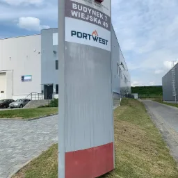 Pylon reklamowy z nazwą Alliance Silesia Logistics Center i Portwest na tle budynku magazynowego w pochmurny dzień.