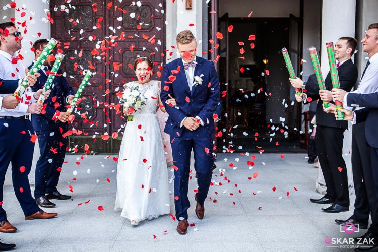 Uśmiechnięta para młoda wychodzi z kościoła, obsypana konfetti w kształcie płatków róż, otoczona przez gości z tubami konfetti.