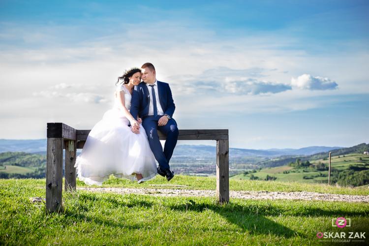 Para młoda siedząca na drewnianej ławce na wzgórzu z widokiem na krajobraz, ona w białej sukni ślubnej, on w granatowym garniturze, trzymają się za ręce, delikatne oświetlenie, błękitne niebo...