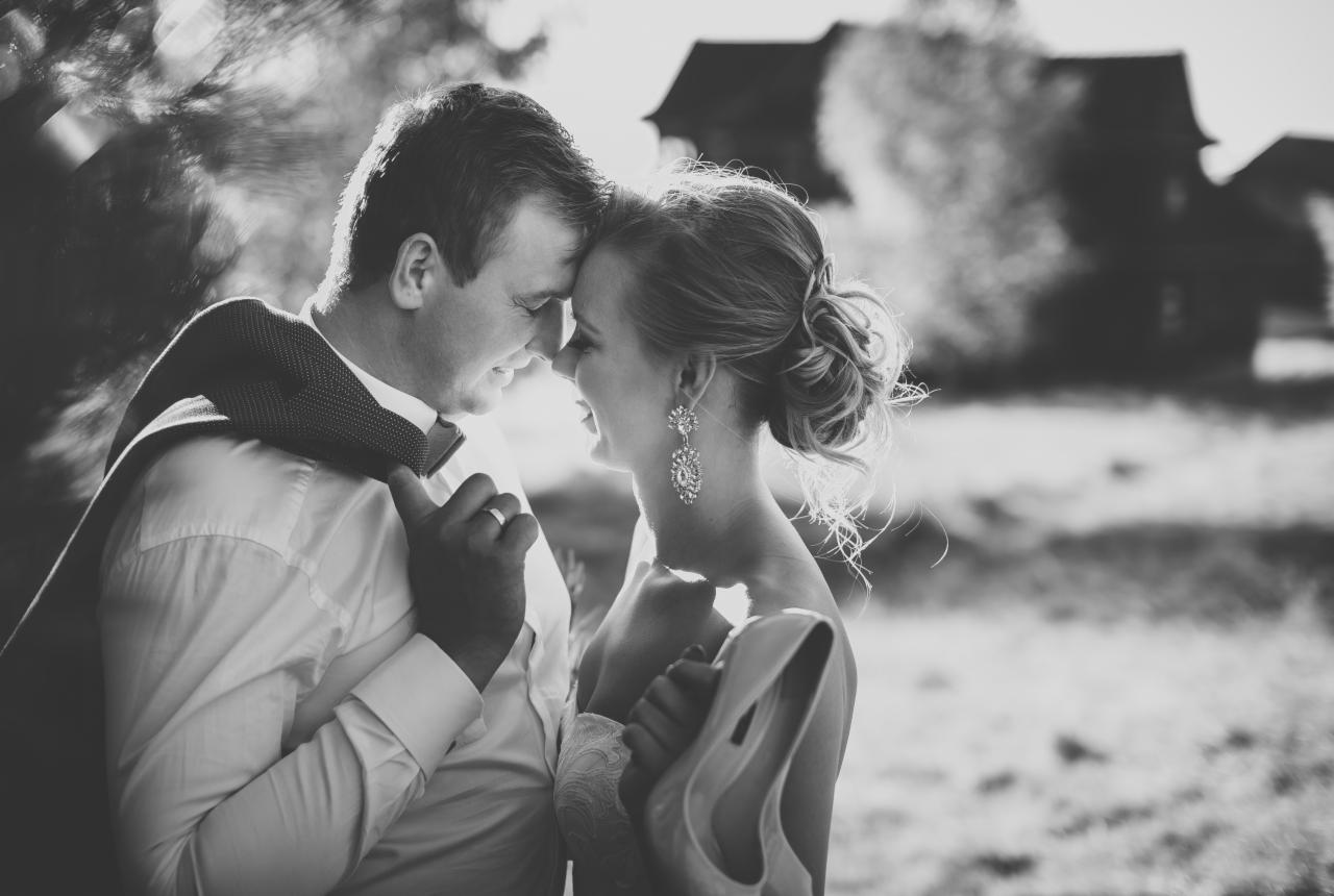 Czarno-białe zdjęcie pary młodej, zbliżenie twarzy, ona trzyma buty, on ma marynarkę na ramieniu, w tle wiejskie zabudowania.