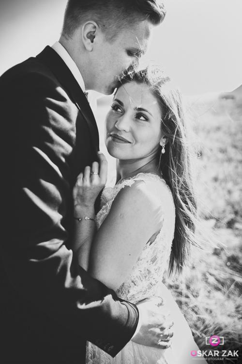 Czarno-białe zdjęcie pary w Nowym Sączu: mężczyzna w garniturze całuje kobietę w koronkowej sukni w czoło, ona spogląda w górę z uśmiechem, w tle łąka.