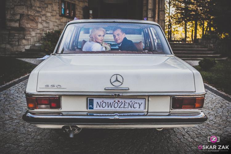Młoda para w białym Mercedesie 220 z tablicą rejestracyjną 'Nowożeńcy', uchwycona z tyłu na tle kamiennego budynku i schodów.