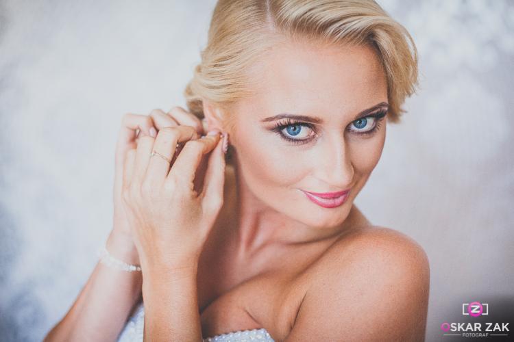 Portret kobiety z blond włosami, w trakcie zakładania kolczyka, z delikatnym makijażem i jasnymi niebieskimi oczami, uśmiechająca się do obiektywu, z widocznym logo fotografa w prawym dolnym rogu.