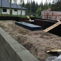 Ruthenia-Group Sp.z o.o - Fundamenty domu w trakcie budowy: betonowe ławy z wystającymi prętami zbrojeniowymi, częściowo zalane, otoczone szalunkiem i ziemią, w tle cegły i drzewa.