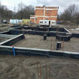 Ruthenia-Group Sp.z o.o - Fundamenty domu jednorodzinnego z widoczną izolacją poziomą i pionową, wystające pręty zbrojeniowe, w tle budynek w stanie surowym otwartym i przyczepy kempingowe.